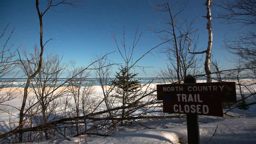 A serene nighttime timelapse captures the star-filled sky above a snowy, frozen lakeshore with a 'Trail Closed' sign in the foreground