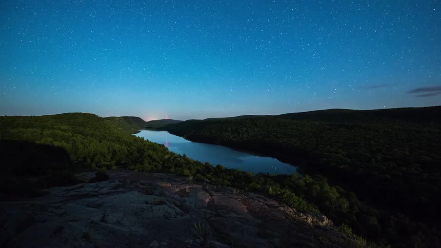 A beautiful day-to-night timelapse from a rocky overlook shows the stars emerging over a serene mountain lake nestled in a lush forest valley