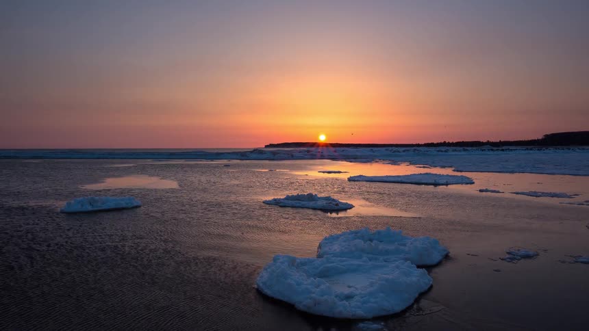 A beautiful timelapse captures the sun rising over a tranquil, icy shoreline, casting a golden glow across the calm water