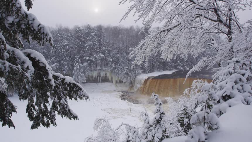 Snow falls gently over a wide, tannic waterfall flowing through a frozen forest landscape