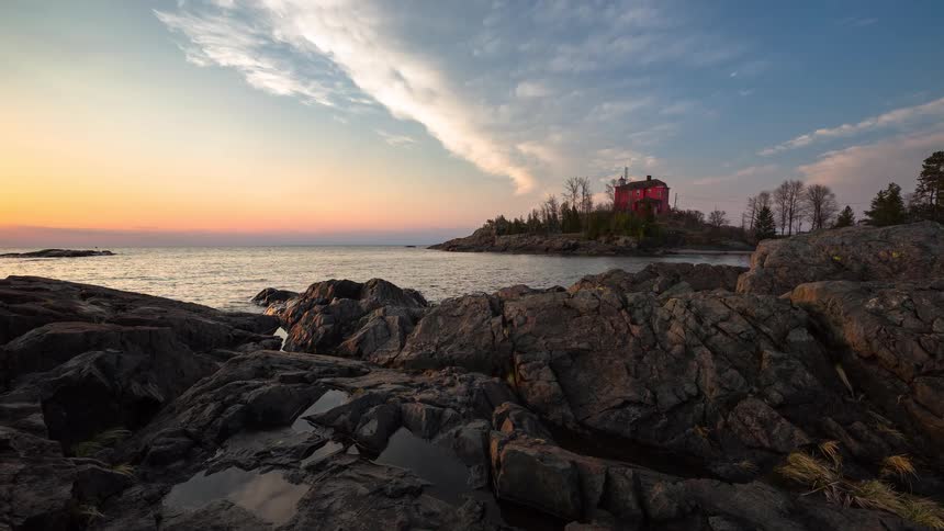 A scenic timelapse captures the changing light of sunset over a tranquil lake, highlighting a red house on a rocky peninsula