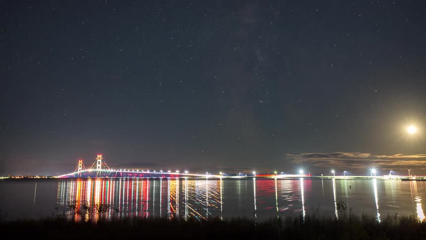 A night time-lapse captures the stars and a bright moon moving over the illuminated Mackinac Bridge as its lights reflect on the water before fog obscures the view