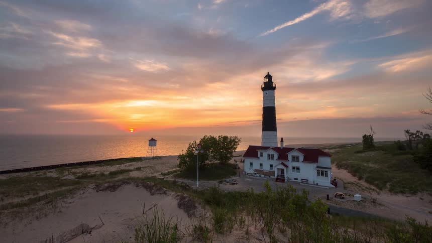 A stunning time-lapse captures the sun setting over the ocean behind the historic Big Sable Point Lighthouse on a sandy beach