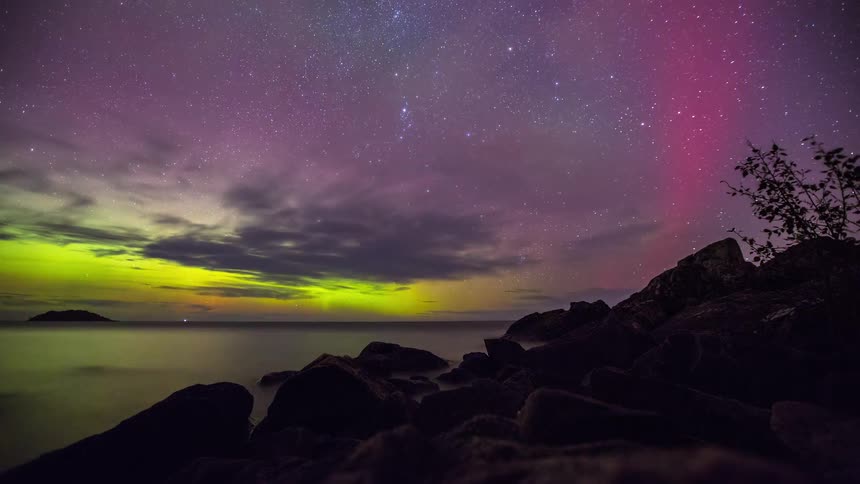 A stunning timelapse shows the vibrant green and purple Aurora Borealis dancing across the starry night sky over a calm lake with a rocky shore