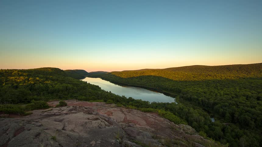 A scenic time-lapse captures the sunset over a lake in a lush green valley, with the sky changing from golden hour to a vibrant pink and purple twilight