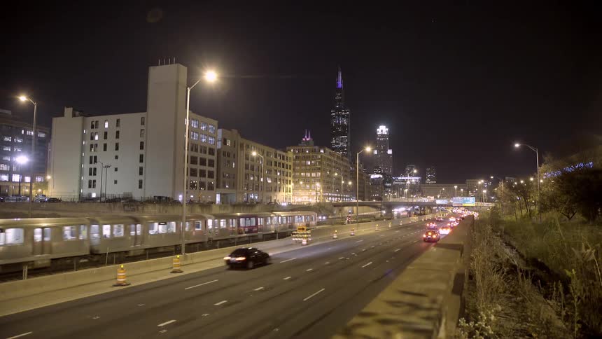 A dynamic nighttime view of Chicago shows cars on the highway and a train passing by with the iconic skyline in the background