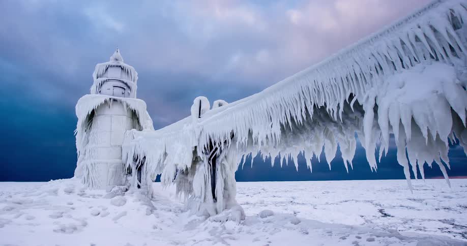 A dramatic winter landscape features a lighthouse and pier completely frozen and covered in long icicles under a stormy sky
