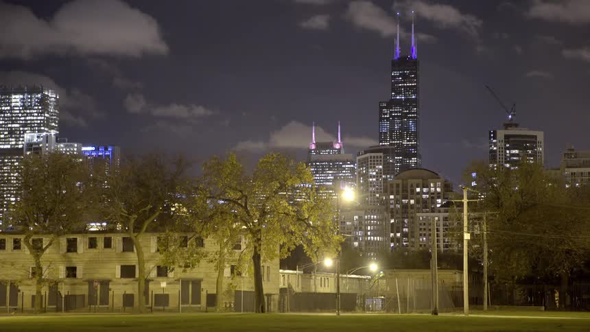 A police car drives through a city park at night with the iconic Chicago skyline and Willis Tower illuminated in the background
