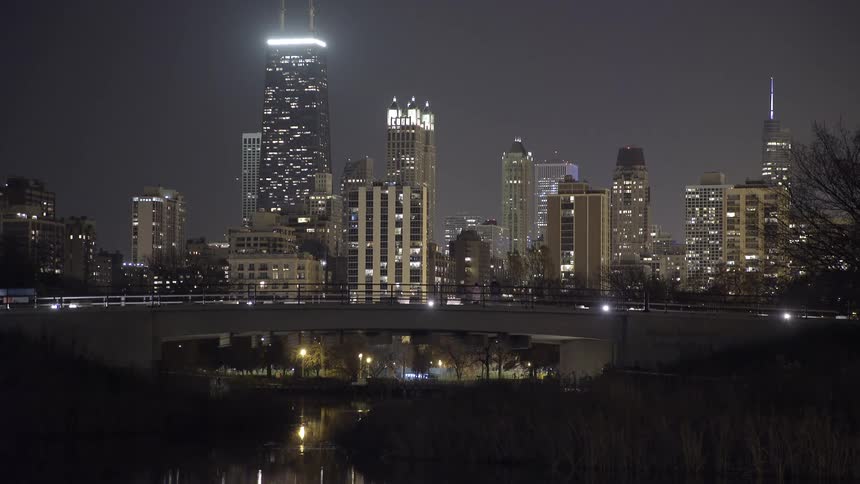 The magnificent Chicago skyline, featuring the John Hancock Center, is illuminated at night over a park and a bridge with passing cars