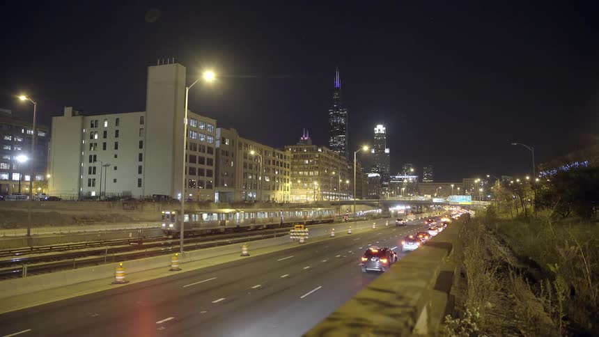 A commuter train and cars travel along a busy Chicago expressway at night with the Willis Tower in the skyline