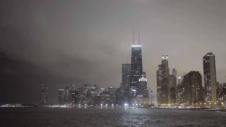 The Chicago skyline, featuring the John Hancock Center, glows at night under a cloudy sky as seen from across Lake Michigan
