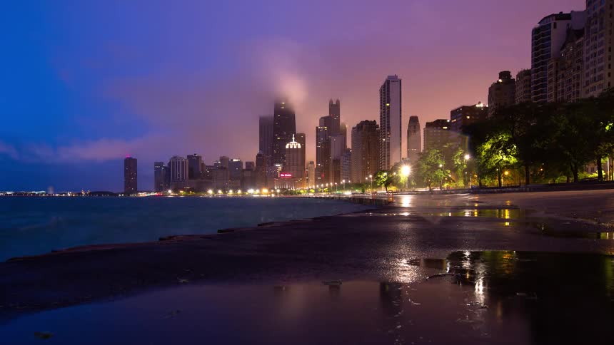 A dramatic time-lapse of a thunderstorm rolling over the Chicago skyline at night, with lights reflecting on the wet lakefront pavement