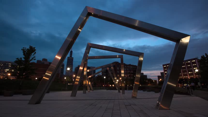 A time-lapse of a modern steel sculpture in a city park as the sun rises over the Chicago skyline in the background