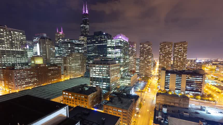 A vibrant time-lapse captures the bustling energy of the Chicago skyline at night from a high-angle view, with light trails from traffic below
