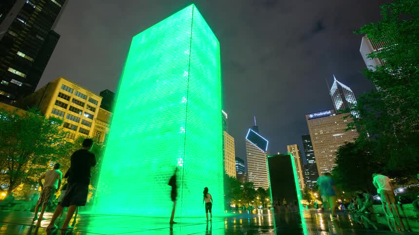 A vibrant night time-lapse shows people playing in the green-lit Crown Fountain with the Chicago skyline in the background