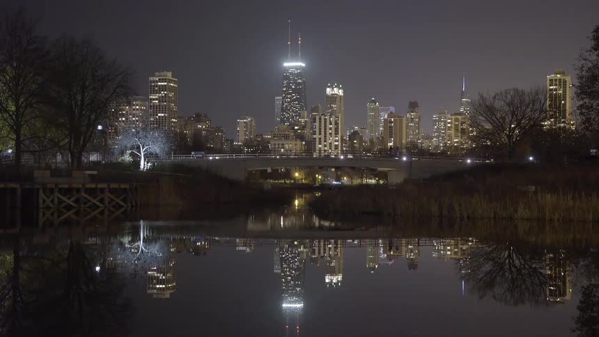 A serene nighttime view of the Chicago skyline, including the John Hancock Center, perfectly reflected in the tranquil waters of a park