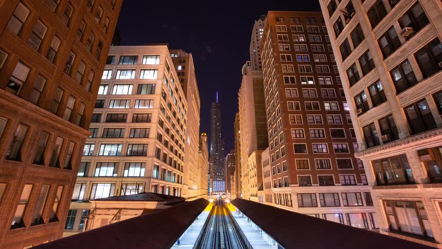 A captivating nighttime timelapse shows the famous 'L' train speeding through a canyon of illuminated skyscrapers in downtown Chicago