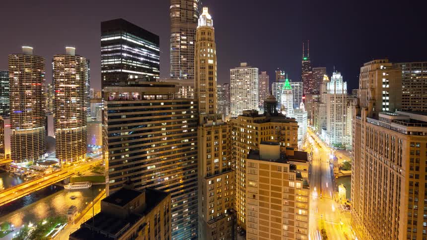 An elevated timelapse captures the vibrant energy of downtown Chicago at night, with light trails from cars and boats weaving through the illuminated cityscape