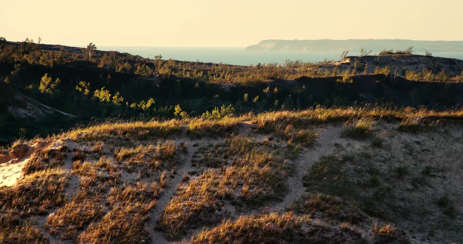 A tranquil view from a grassy sand dune shows a hiking path overlooking the coast during a warm sunset