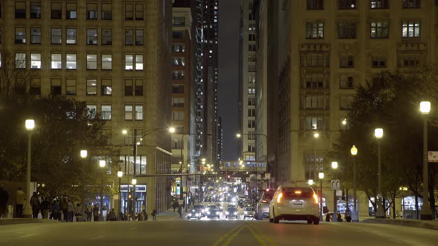 A slow-motion nighttime view of a bustling city street, with cars driving toward the camera between tall buildings