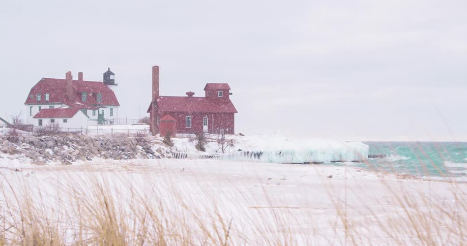 A historic lighthouse and its outbuildings are covered in snow during a winter storm on the icy shores of a large lake