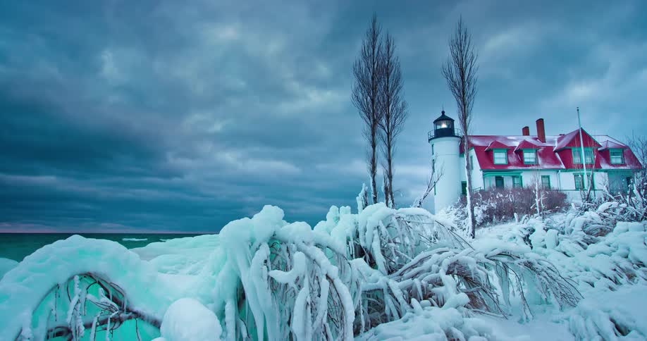 A picturesque lighthouse stands guard over a frozen, ice-covered shoreline as stormy winter clouds drift across the sky