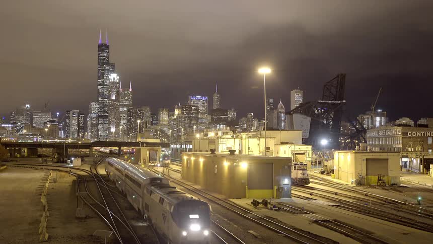 An Amtrak passenger train travels through a rail yard at night with the illuminated Chicago skyline in the background