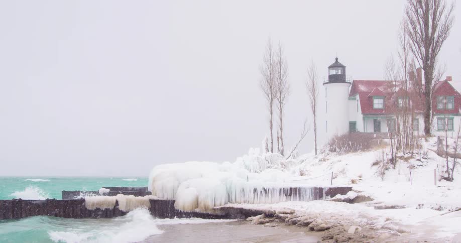 A snow-covered lighthouse on an icy shoreline endures crashing waves during a winter storm in slow motion