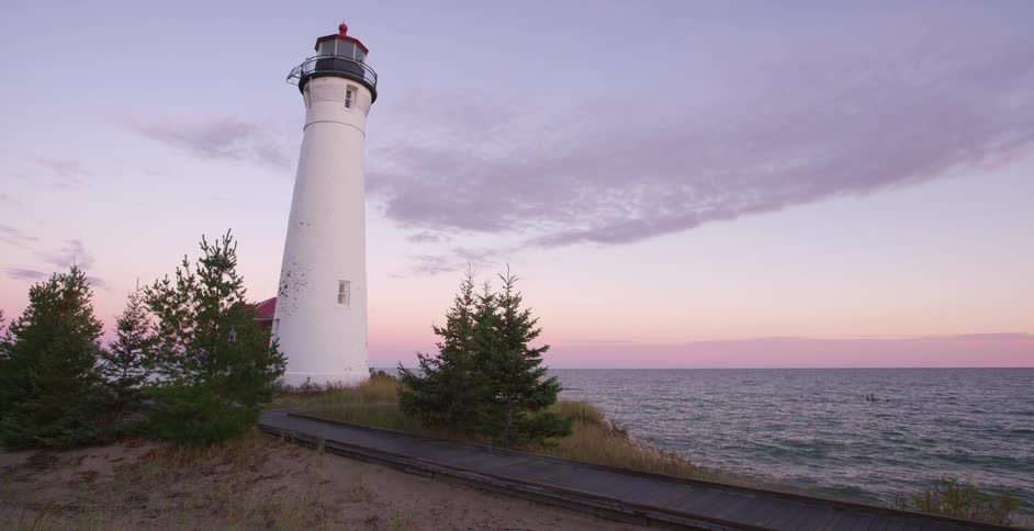 A picturesque white lighthouse stands on a grassy dune beside the ocean during a peaceful pastel-colored sunset