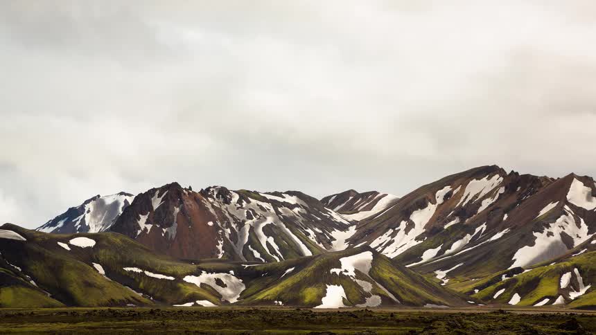 A timelapse of clouds moving swiftly over a rugged, multicolored mountain range partially covered in snow