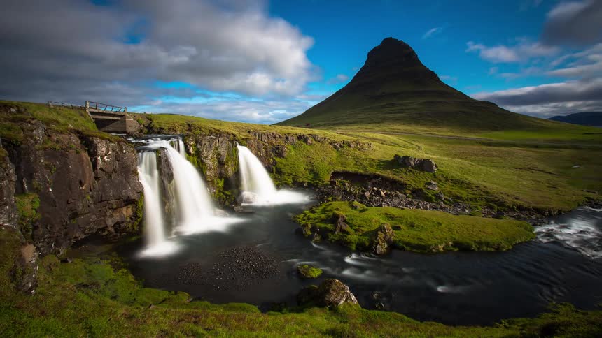 A stunning timelapse of the iconic Kirkjufell mountain and Kirkjufellsfoss waterfall in Iceland, with clouds moving across a bright blue sky