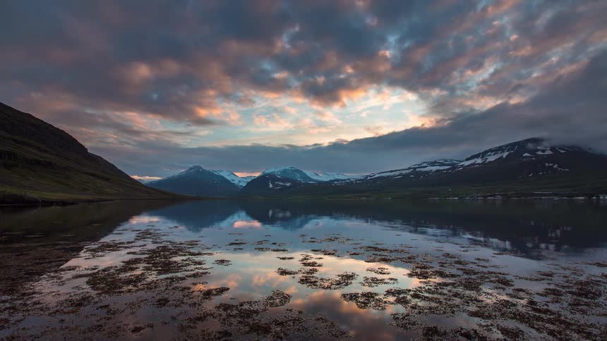 A majestic time-lapse captures a dramatic sunset and clouds reflecting in the still waters of a fjord surrounded by snow-capped mountains