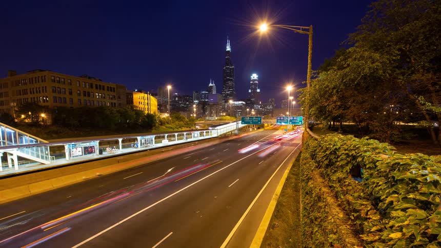 A long exposure time-lapse captures the vibrant light trails from vehicle traffic on a busy Chicago highway with the illuminated city skyline in the distance