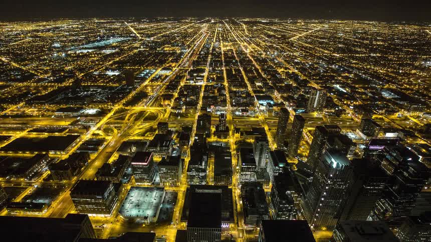 An expansive aerial time-lapse captures the vibrant energy of a massive city at night, with streaks of light from traffic flowing through the illuminated grid