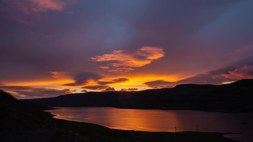 A beautiful timelapse shows a fiery orange and purple sunset reflected in the still waters of a mountain fjord