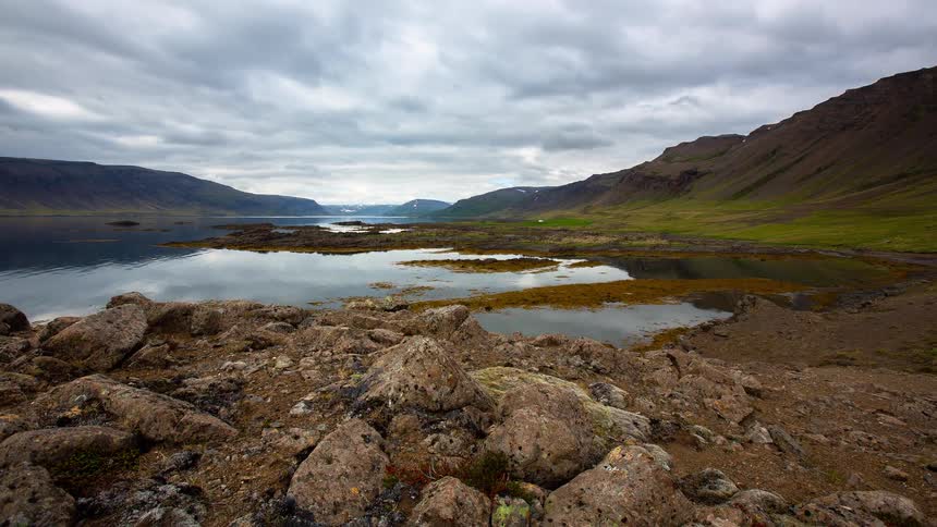 Timelapse of dramatic clouds passing over a serene fjord in Iceland, viewed from a rocky shoreline with green mountains in the distance