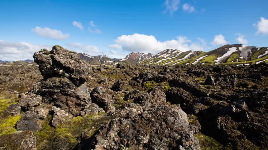 A stunning timelapse of clouds and shadows moving across a mossy volcanic lava field in front of colorful, snow-capped mountains in the Icelandic highlands
