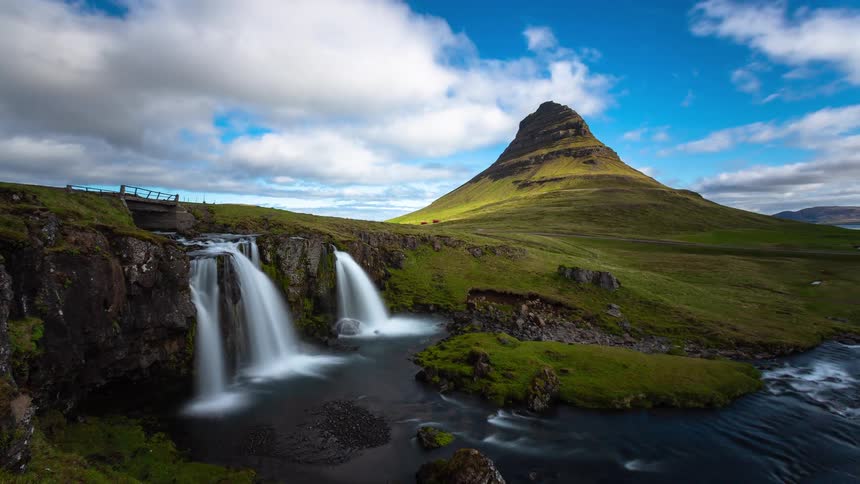 A majestic timelapse of the iconic Kirkjufellsfoss waterfall and Kirkjufell mountain in Iceland as clouds drift across a vibrant blue sky