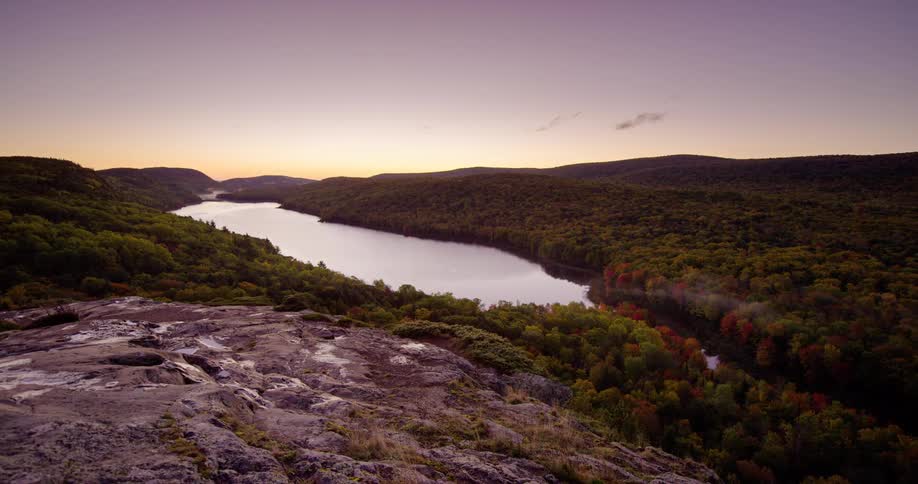 A serene timelapse from a rocky mountain overlook captures a tranquil lake surrounded by a dense forest with vibrant autumn foliage at dawn