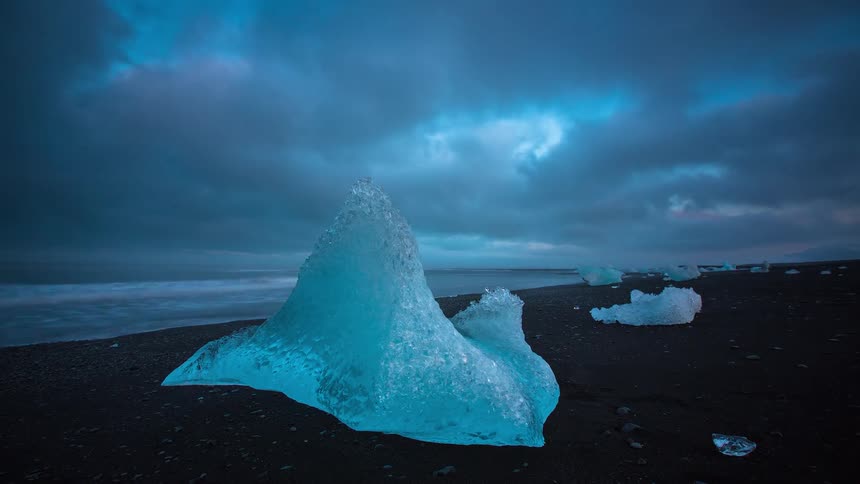 Timelapse of a glacial ice chunk on Iceland's black sand beach as a colorful sunset fades to a dark, cloudy night