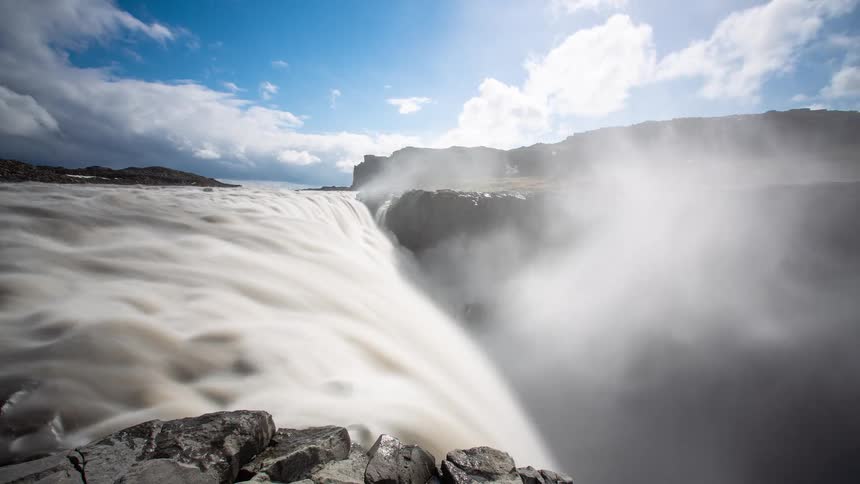 Long exposure timelapse captures the silky smooth flow of a massive waterfall plunging into a misty canyon under a dynamic cloudy sky