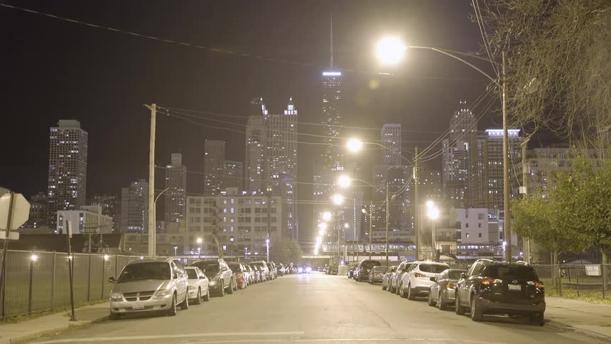 A car slowly drives toward the camera on a quiet city street with the hazy Chicago skyline illuminated in the background at night