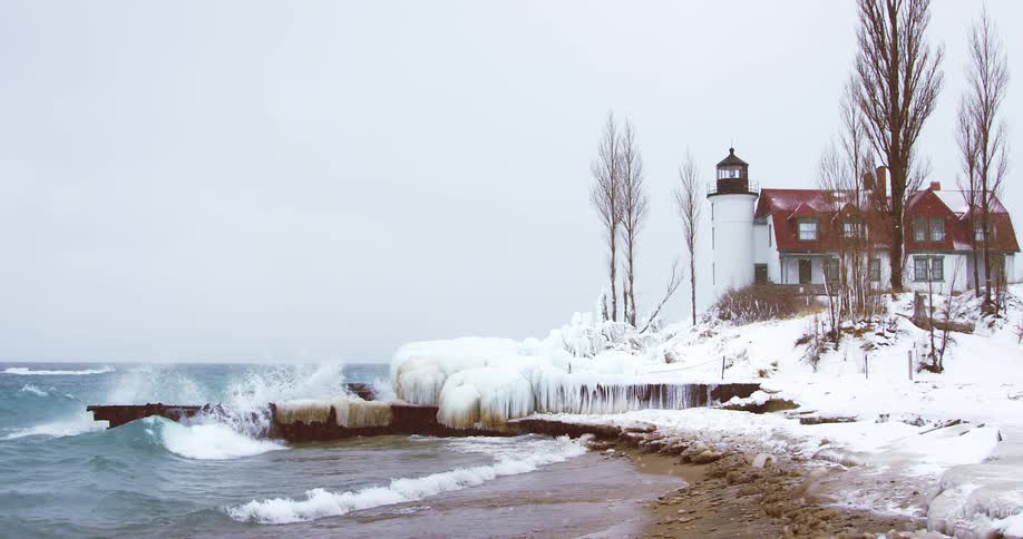 A lighthouse stands on a snowy, ice-covered shore as large waves crash against the pier in slow motion during a winter storm