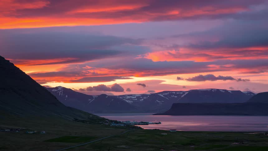 A time-lapse captures a fiery red and purple sunset with moving clouds over a tranquil Icelandic fjord and its distant, snow-capped mountains