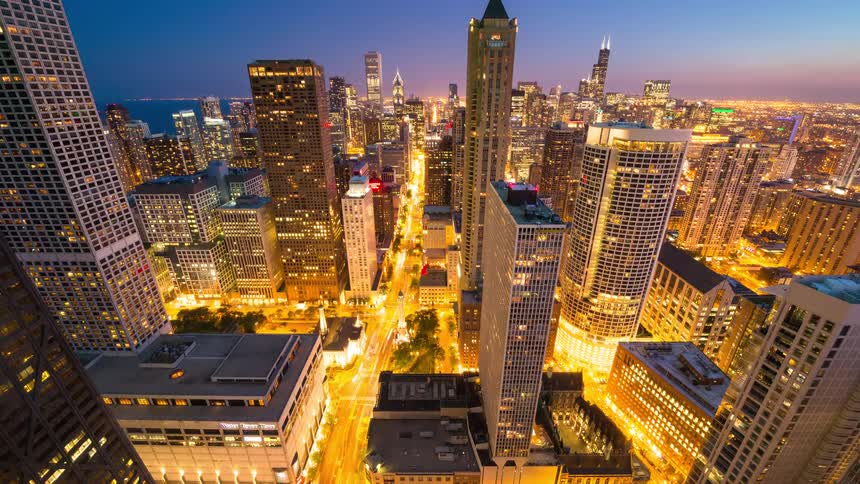 A high-angle timelapse captures the transition from a sunlit afternoon to a brightly lit evening in downtown Chicago, with traffic creating light trails on the streets below