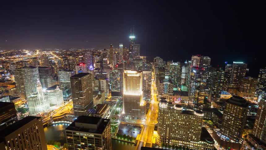 A wide aerial time-lapse of the Chicago skyline at night with traffic and boats creating vibrant light trails through the bustling city