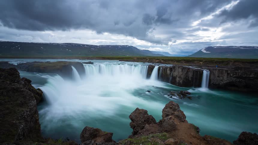 A long exposure timelapse captures the powerful flow of the Goðafoss waterfall in Iceland under a dramatic, cloudy sky