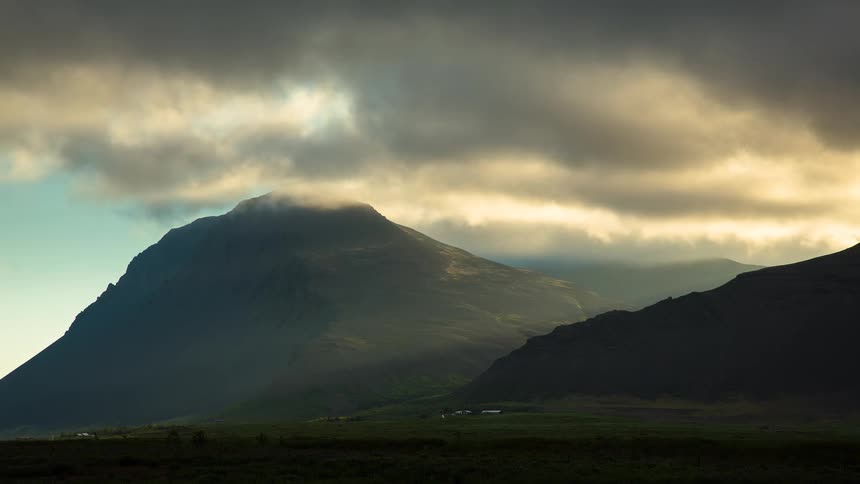 Dramatic dark clouds drift over a majestic mountain peak illuminated by warm, golden light