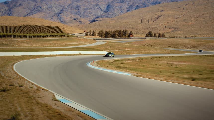 A sleek sports car with green accents speeds around a winding corner on a professional racetrack in an arid landscape