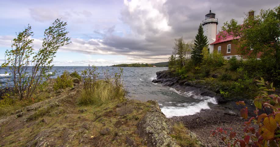 A historic lighthouse stands guard over a rocky shoreline as gentle waves roll in under a moody, cloud-filled sky in autumn